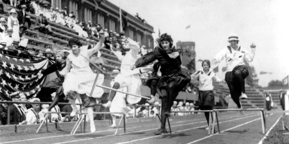 Women compete in a hurdle race on a track with flag bunting in the background. 